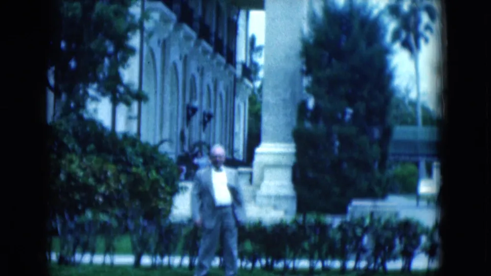 1975 Florida 8mm frame showing a suited man on grass before hedges, palms, arched windows, and a classical facade.