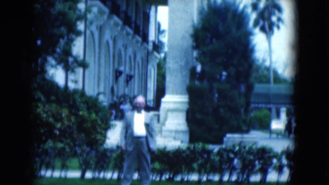 1975 Florida home-movie frame of a man in a light suit near manicured hedges, palms, and a formal building exterior.