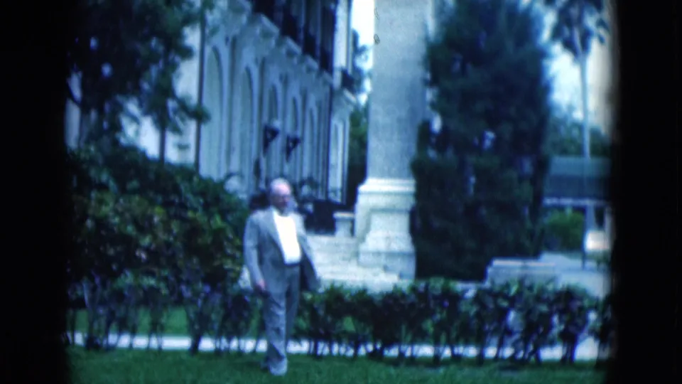 1975 Florida 8mm still of a suited man walking on a lawn before low hedges and a classical building with arched windows.