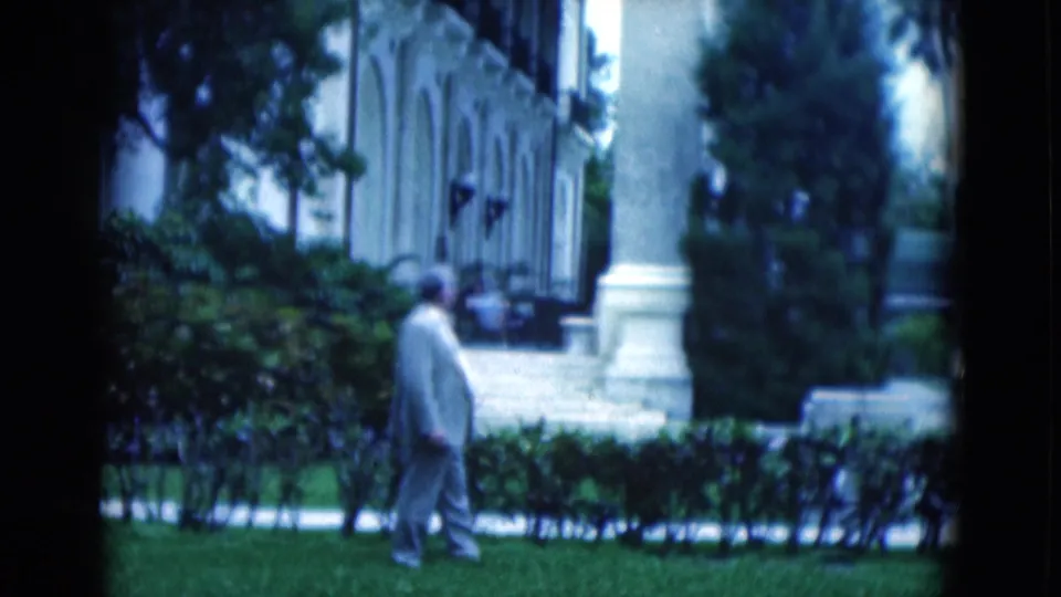 1975 Florida home-movie frame with the suited man in profile, walking past hedges near palms and a columned building exterior.