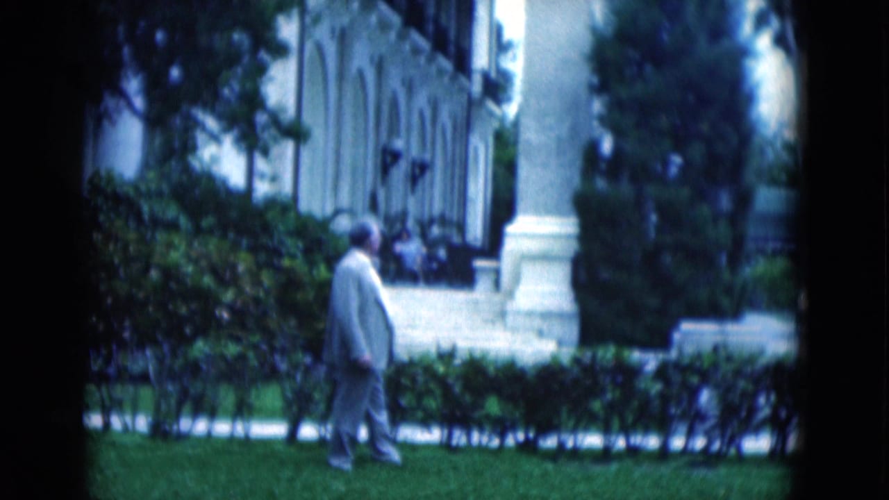 1975 Florida 8mm frame showing an adult man on grass beside hedges and a white building with columns.