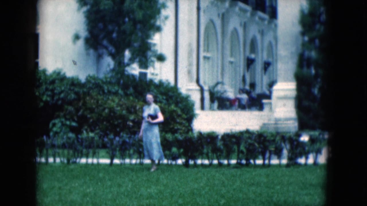 Woman in a light dress walking across a lawn in front of hedges and arched windows.