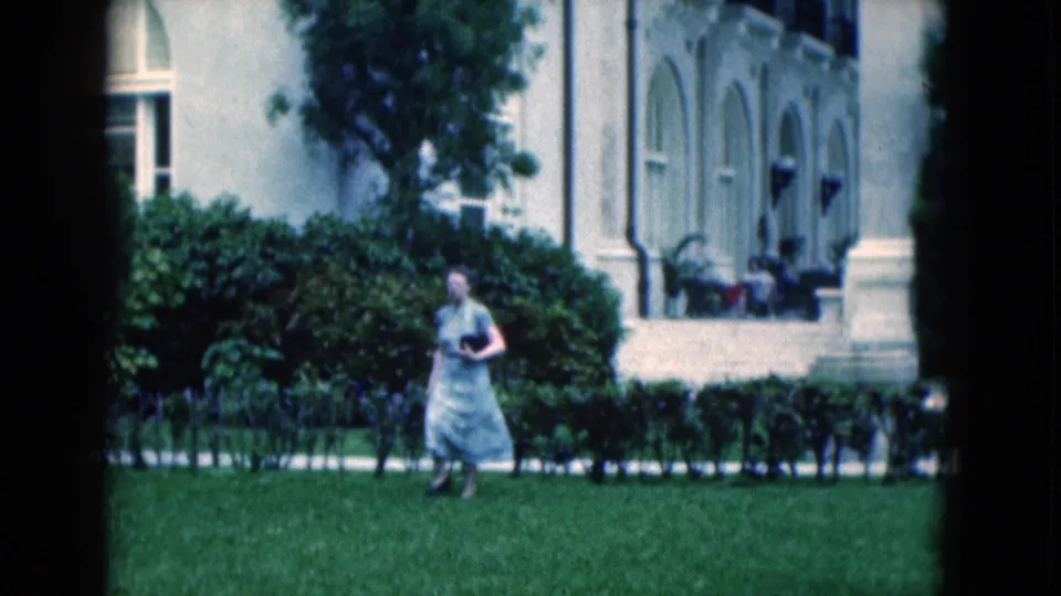 1975 Florida home-movie frame showing a woman in a pale dress walking past hedges and a white columned building.