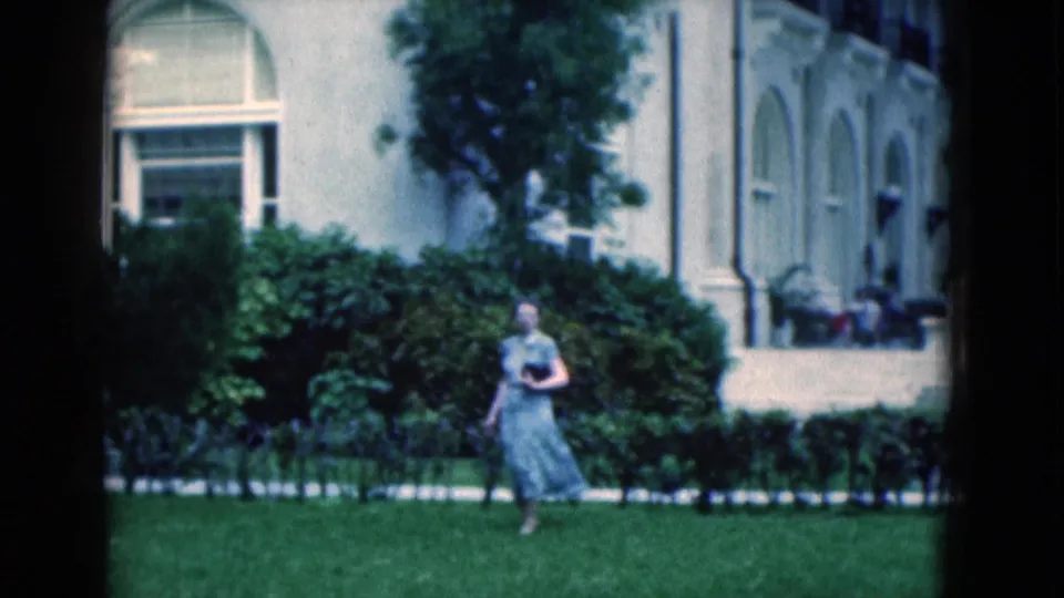 1975 Florida 8mm frame of a woman crossing a manicured lawn in front of a white building and shrubs.