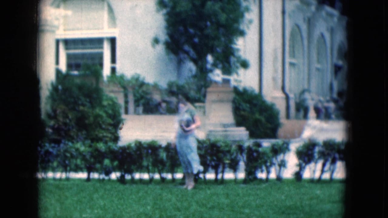 Woman walking beside hedges with a white arched building behind her.