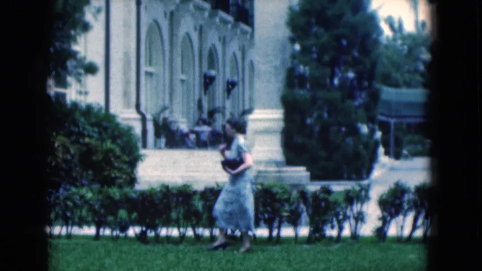 A soft 8mm view shows the woman near the foreground hedge, with steps, arched windows, and outdoor seating behind her.
