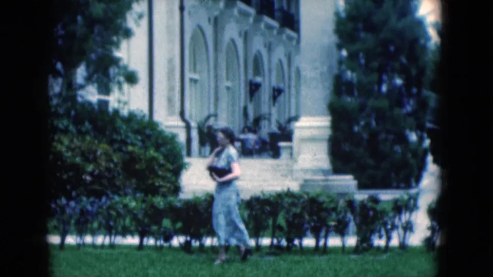 The final frame keeps the woman in profile on the lawn, with trimmed hedges, steps, trees, and the white building behind her.
