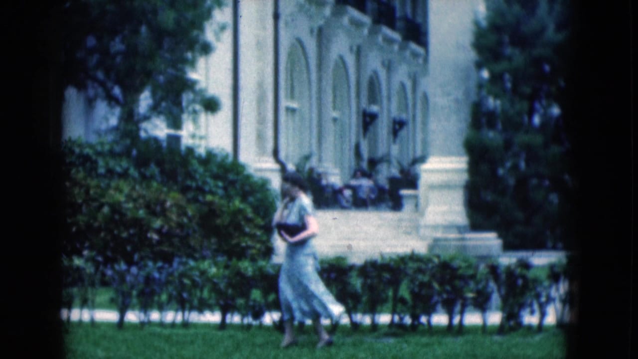 1975 Florida 8mm frame showing a woman in a light dress crossing a manicured lawn before hedges and a formal arched facade.