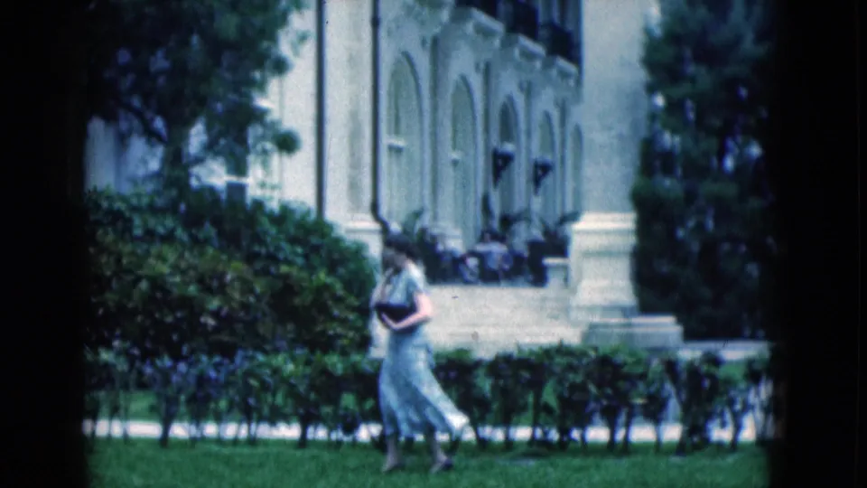 1975 Florida 8mm frame showing a woman in a light dress crossing a manicured lawn before hedges and a formal arched facade.