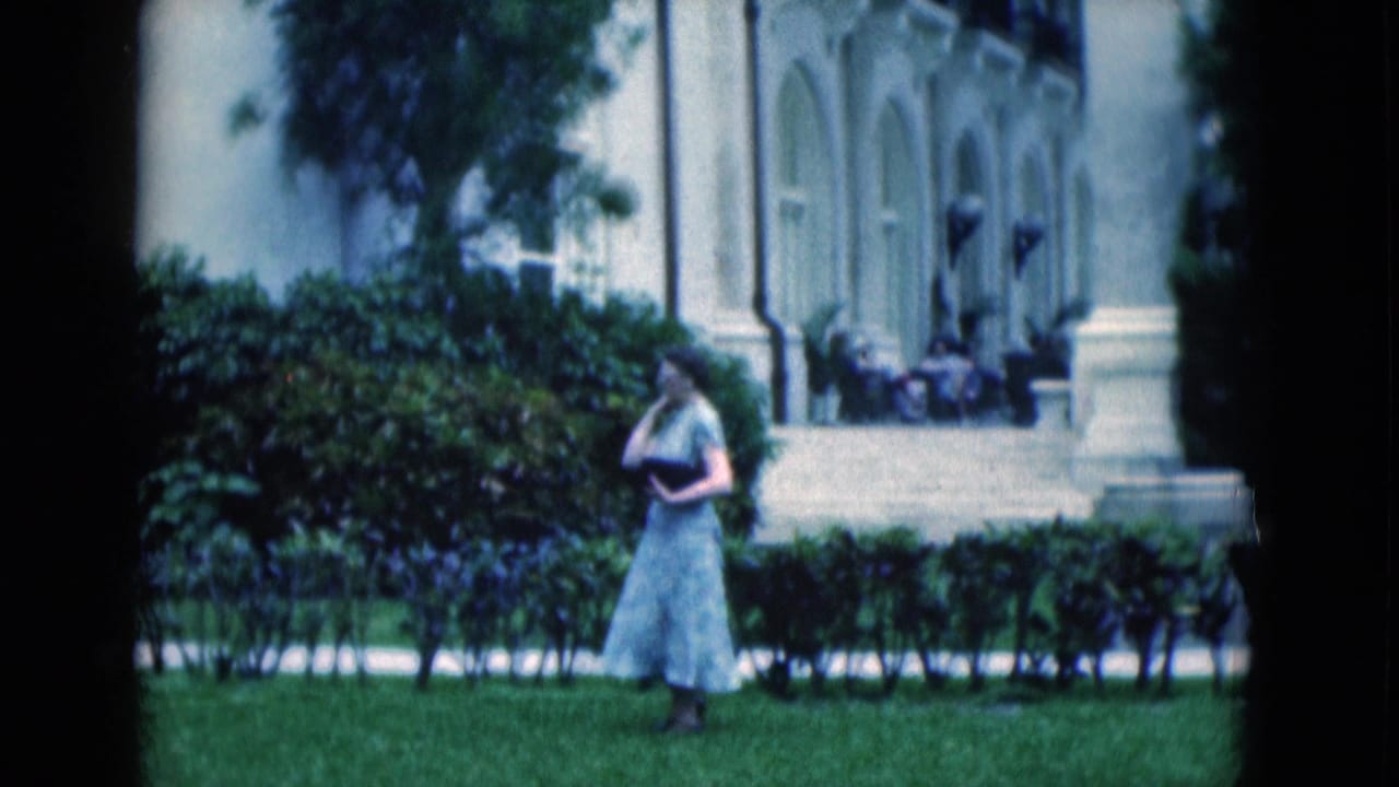 1975 Florida home-movie frame with a woman in a light dress near hedges, backed by a pale building with columns and arched openings.