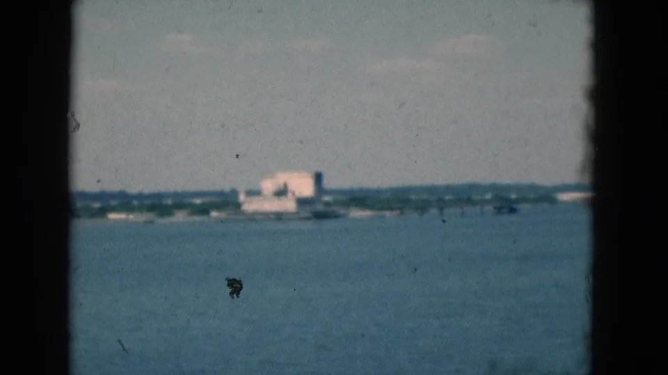 1975 Florida 8mm frame showing a distant pale building across a body of water, with low shoreline, hazy sky, and film specks.