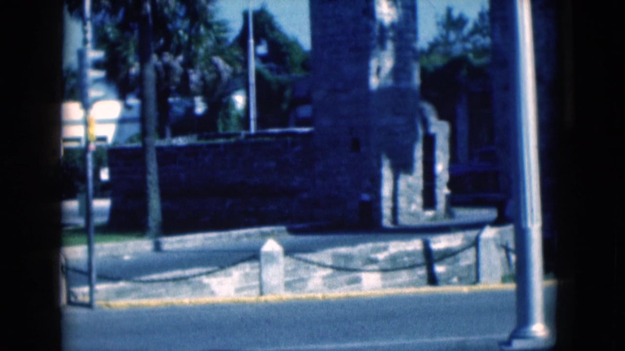 1975 Florida 8mm exterior frame showing a stone structure near a roadside curb, metal poles, trees and a chain barrier.
