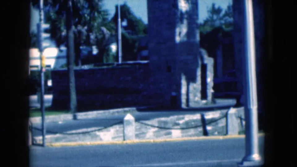 1975 Florida 8mm exterior frame showing a stone structure near a roadside curb, metal poles, trees and a chain barrier.
