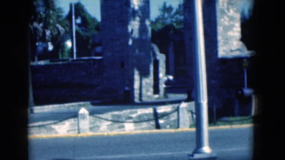 1975 Florida 8mm frame with a blue-tinted roadside view of a stone structure, tall metal pole, curb and chain barrier.