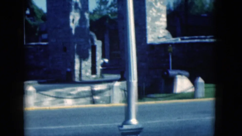 1975 Florida 8mm exterior frame showing a stone structure beside a paved road, with poles, curb, trees and chain barrier visible.