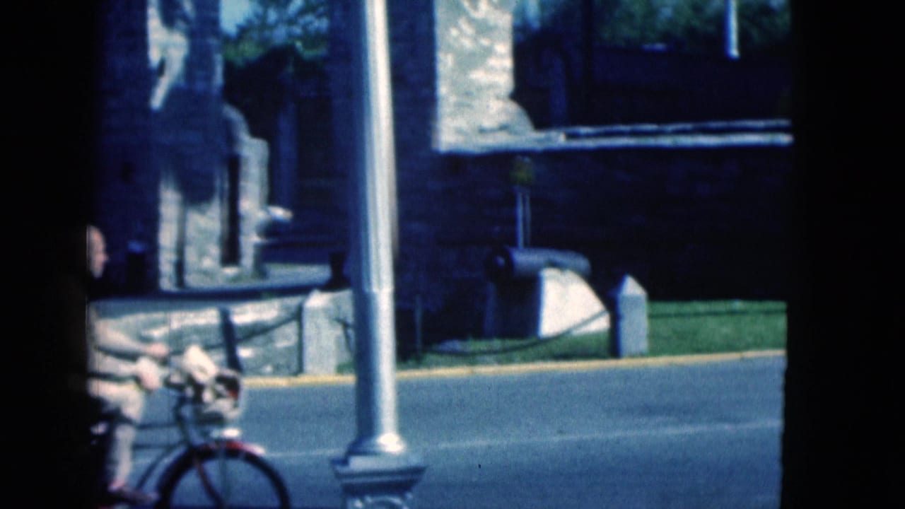 In 1975 Florida home-movie footage, a man on a bicycle passes a stone wall and civic-looking tower area beside a road.