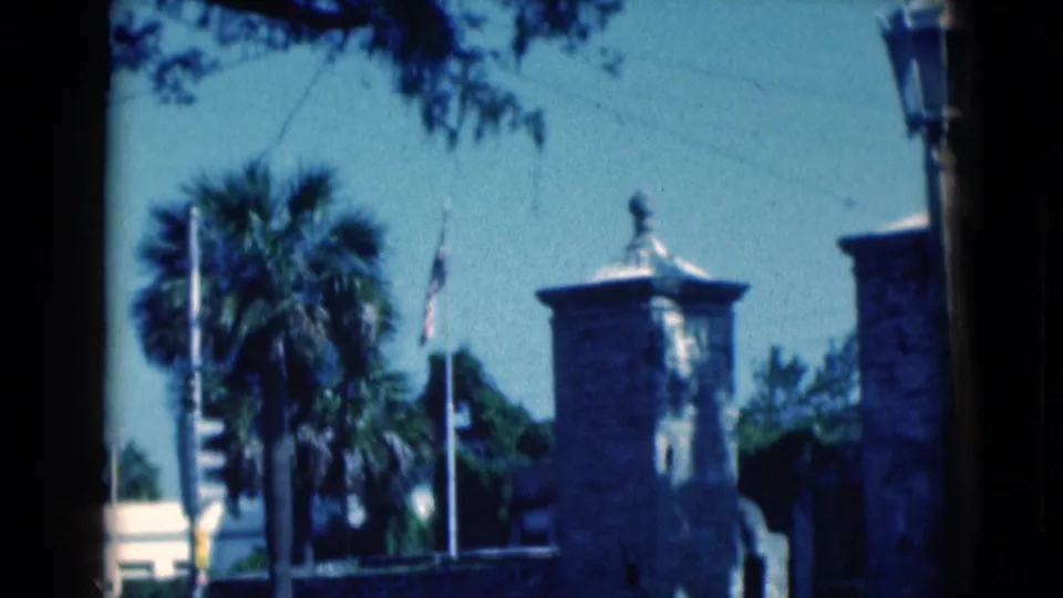 A 1975 Florida frame shows an American flag near a stone tower, palm tree, and white poles under a bright blue sky.