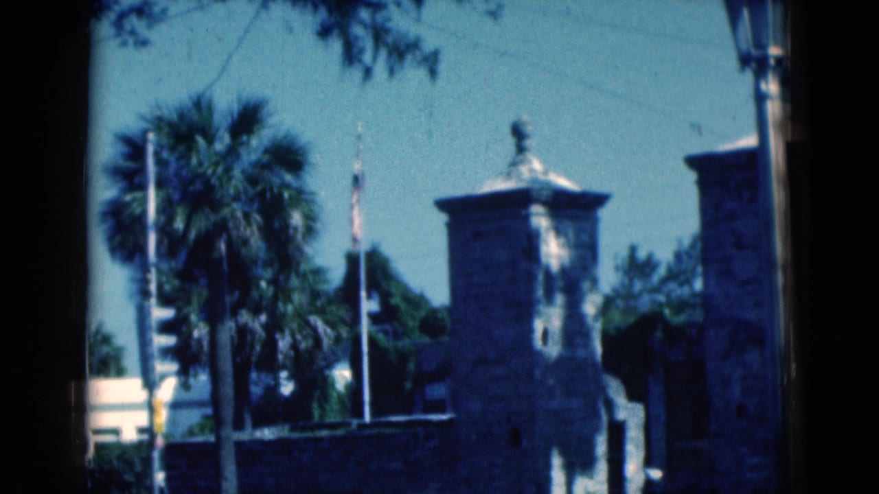 1975 Florida 8mm frame showing historic-looking stone gate towers, palm trees, flags, and a street lamppost in bright sun.