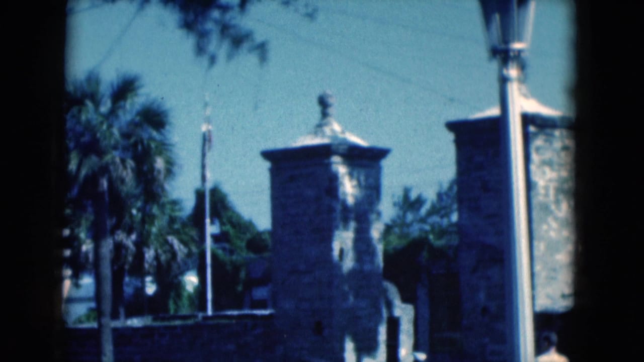 Stone masonry towers, palm tree, flagpole, and lamppost framed in soft daylight.
