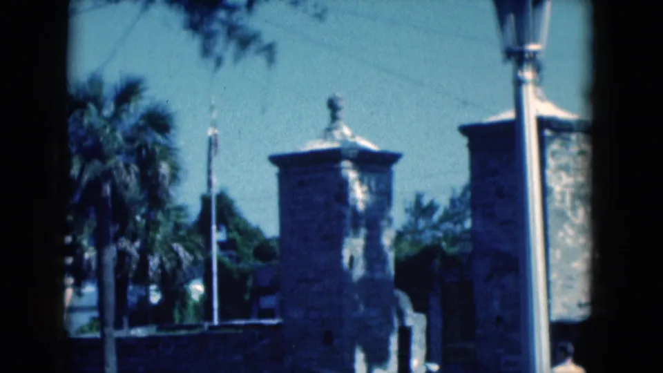 1975 Florida home-movie still with stone gate towers, a palm tree, flags, and a tall lamppost near the street.