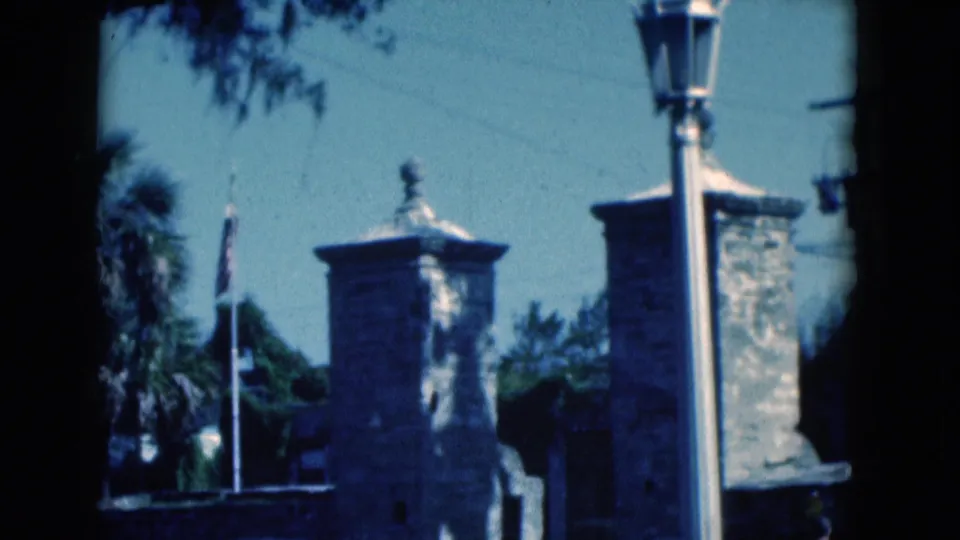 1975 Florida 8mm frame showing stone towers, flags, palm fronds, and a bright lamppost at the edge of the composition.