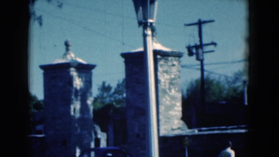 1975 Florida street-side still with stone gate towers, a lamppost, utility pole, overhead wires, and a small distant pedestrian.