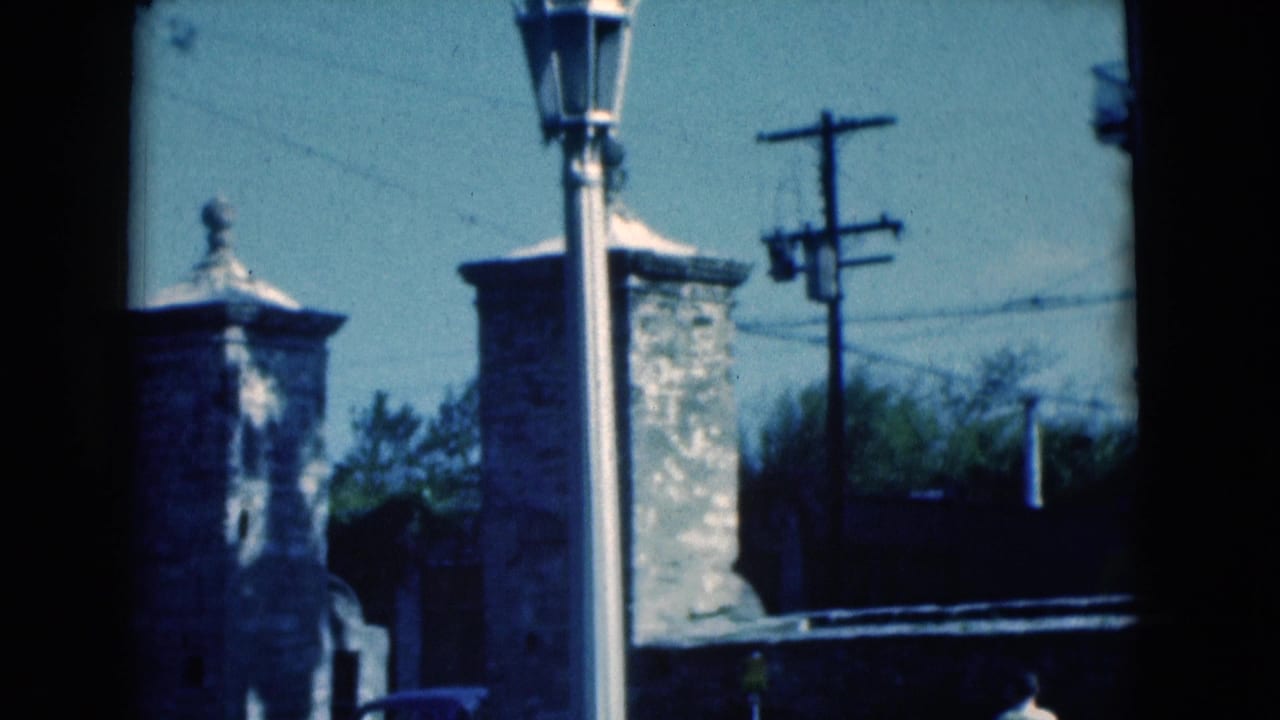 Stone gate pillars, a tall lamp post, utility pole, and trees beside a sunlit road.