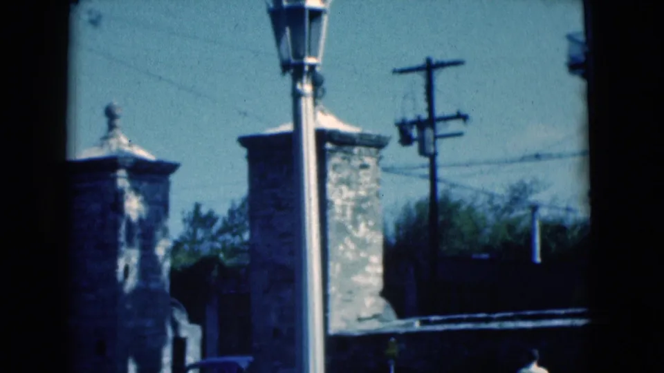 1975 Florida 8mm frame showing stone gate pillars, a tall lamp post, utility wires, and trees near a roadside wall.
