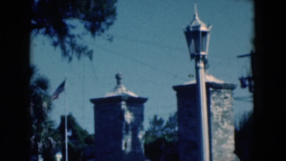 1975 Florida home-movie frame with an American flag, stone pillars, lamp post, trees, and masonry wall near the road.