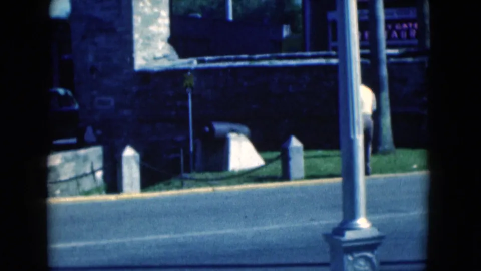 1975 Florida roadside frame showing a lamp post, stone wall, utility pole, grass, road, and a partially visible person near the edge.