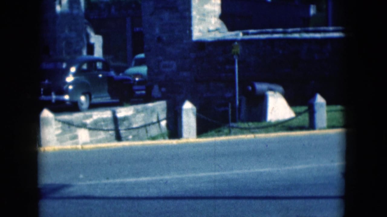 1975 Florida home-movie frame with a rounded sedan beside a stone building and low wall across the street.