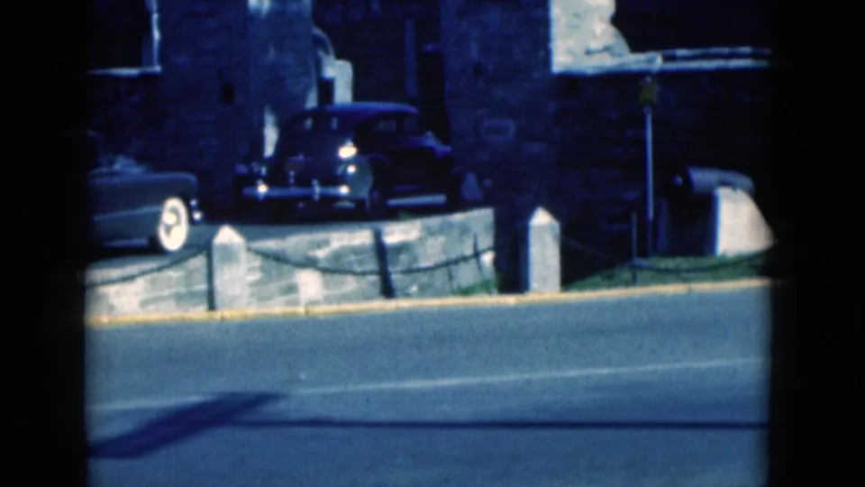 1975 Florida 8mm frame showing classic cars near a stone wall with the road in the foreground.