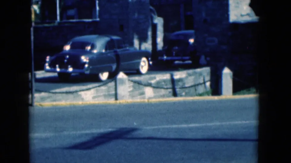 1975 Florida home-movie frame of a dark vintage sedan driving past a stone building and visible cannon display.