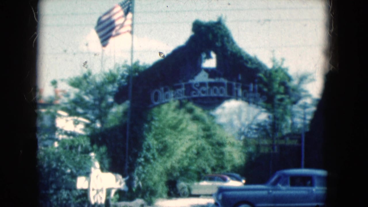 1975 Florida home-movie frame showing the Oldest School House arch, American flag, greenery, and cars near the entrance.