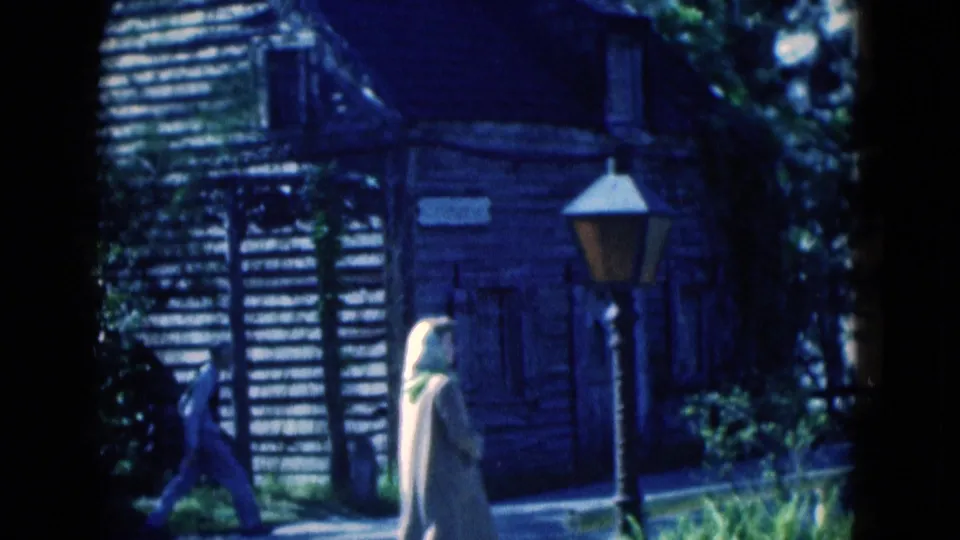 1975 Florida 8mm frame showing a rustic wooden building, lamp post, shaded walkway, and visitors near the Oldest School House site.