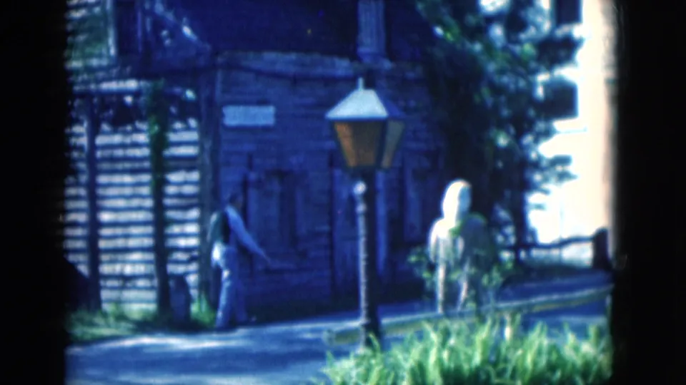1975 Florida home-movie frame of visitors beside the Oldest School House building, with a lamp post, walkway, and sunlit greenery.
