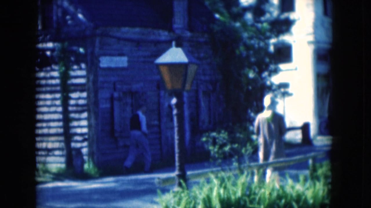 Two pedestrians near a wooden building and street lamp on a shaded street.