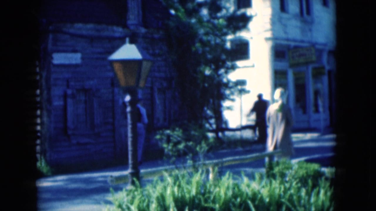 1975 Florida 8mm frame with pedestrians near a wooden streetfront, decorative lamp, shaded foliage, and a bright doorway or road opening.