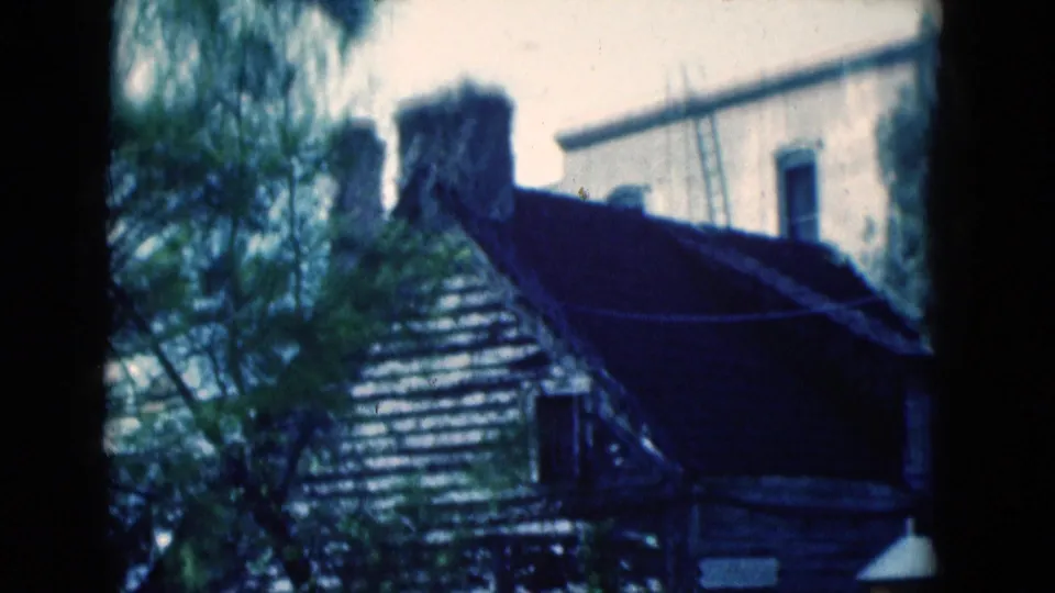 1975 Florida 8mm frame showing a weathered wooden structure, tree branches, roofline, and strong shadow near a decorative lamp edge.