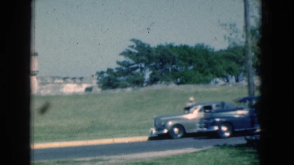 1975 Florida 8mm frame showing a light-colored classic car on a road beside open grass, trees, and a low stone or masonry wall.