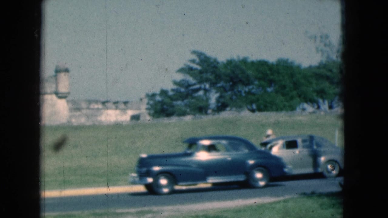 1975 Florida 8mm frame showing blue and silver classic cars driving past a grassy stone fort, viewed through dark window-frame edges.