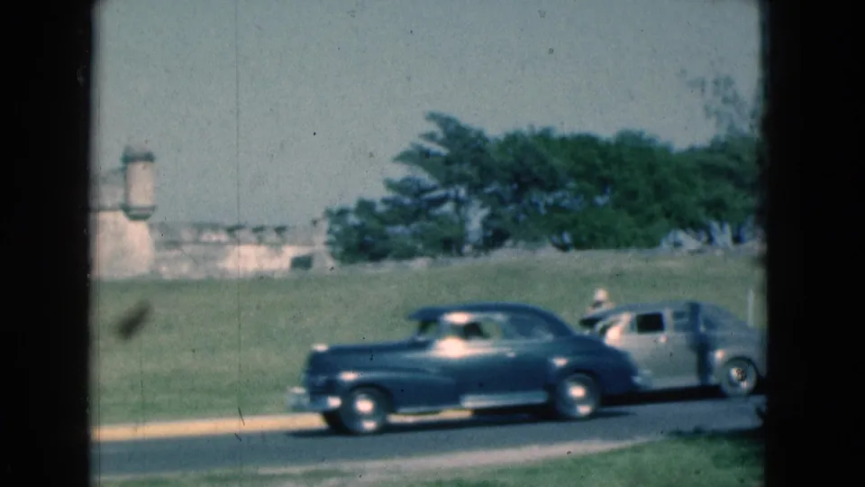 1975 Florida 8mm frame showing blue and silver classic cars driving past a grassy stone fort, viewed through dark window-frame edges.