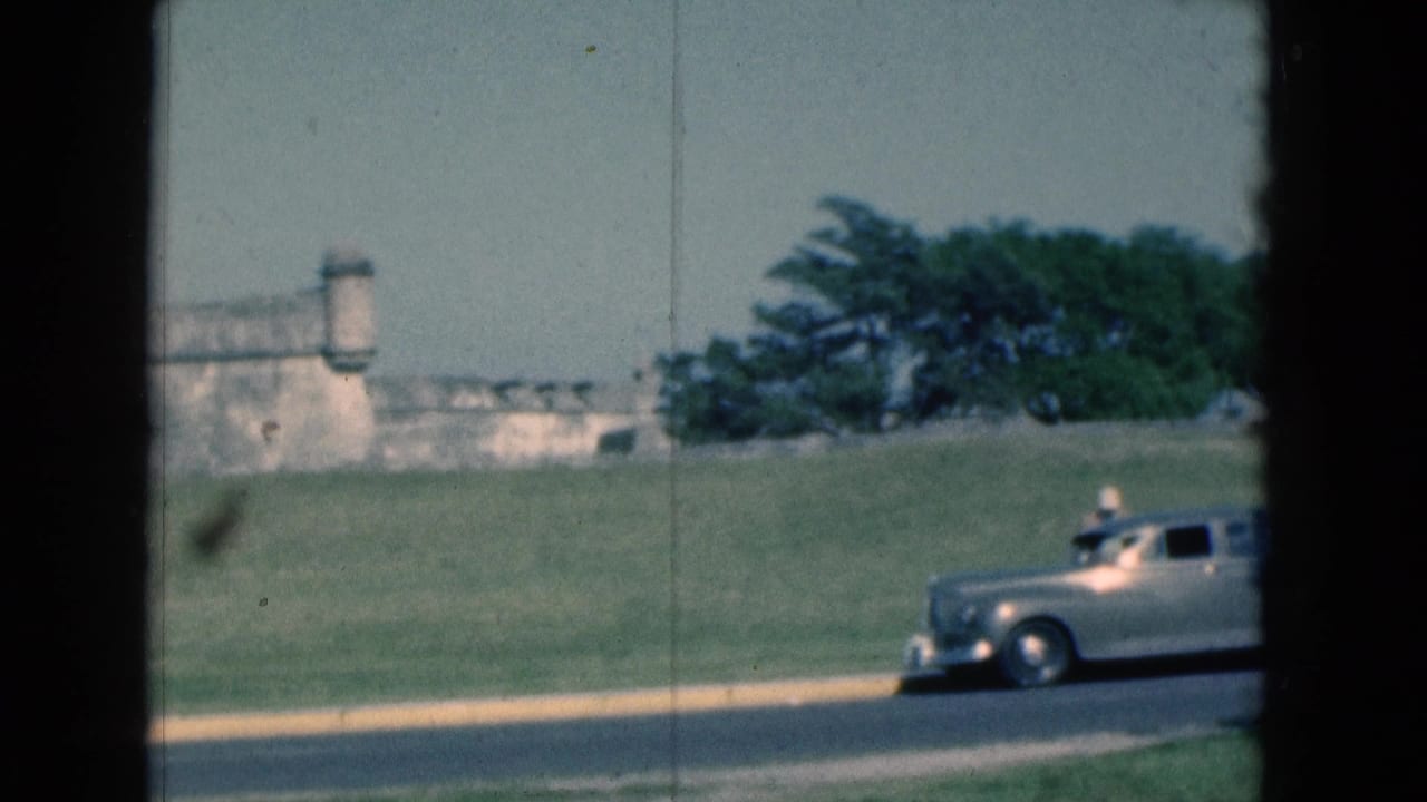 1975 Florida 8mm frame with a silver classic car passing below a weathered stone fort wall and trees on a grassy rise.