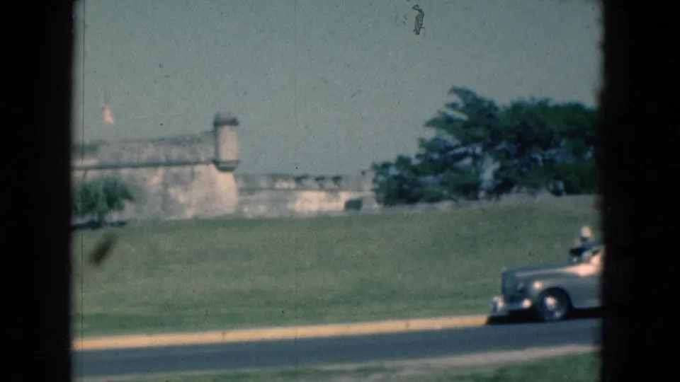 1975 Florida 8mm frame showing the front of a silver classic car at the edge of a road below a stone fort and flag.