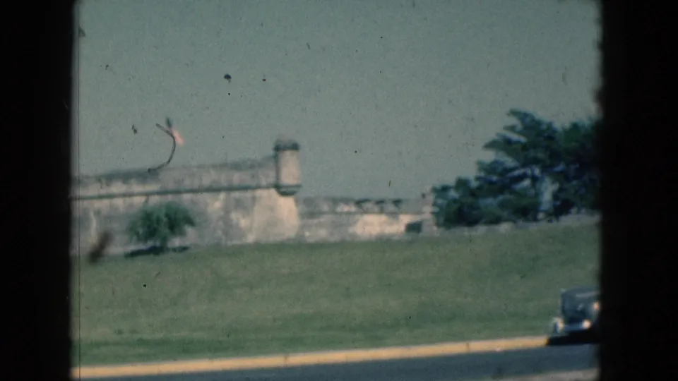 1975 Florida 8mm frame with the stone fort, trees, grassy slope, and the edge of a departing car seen through a dark frame line.
