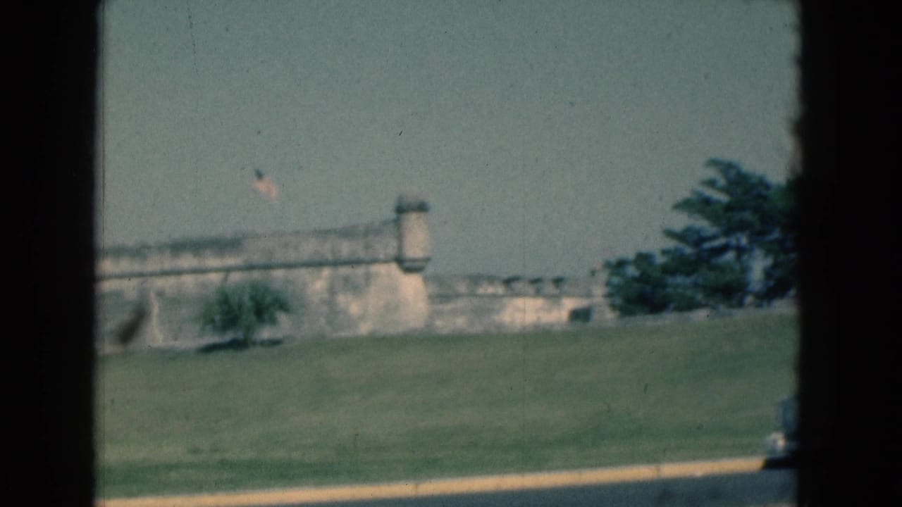 1975 Florida 8mm frame showing a weathered stone fort wall, small tower, flag, lawn, and roadside edge from a moving vehicle.
