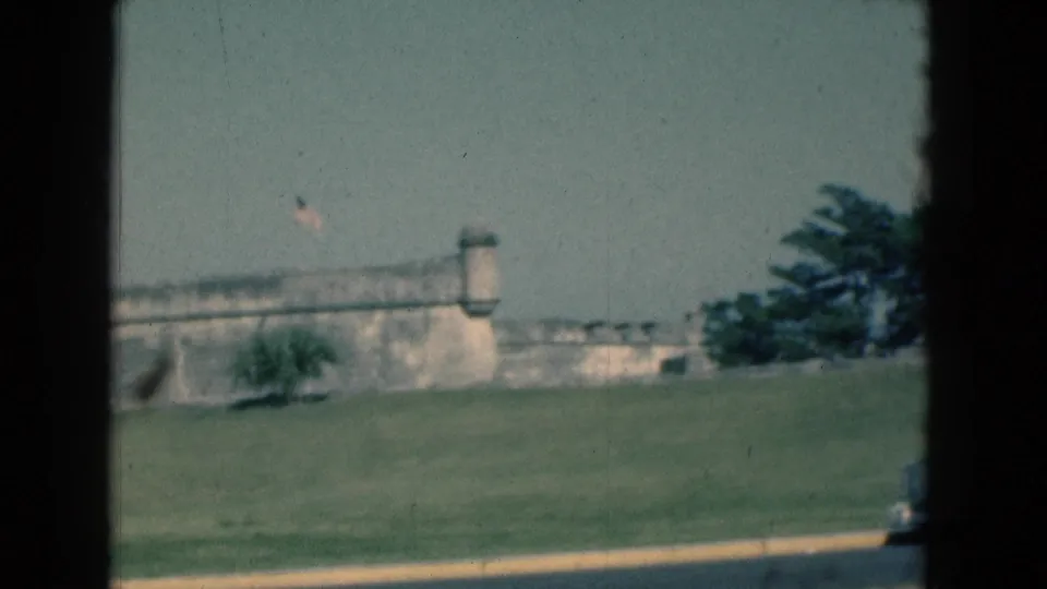 1975 Florida 8mm frame showing a weathered stone fort wall, small tower, flag, lawn, and roadside edge from a moving vehicle.