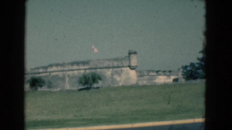1975 Florida home-movie frame with a stone fort wall, flag, lawn, trees, and dark 8mm frame edges.