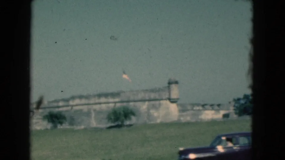 1975 Florida 8mm frame showing the fort wall and flag as a dark sedan appears along the road in the foreground.