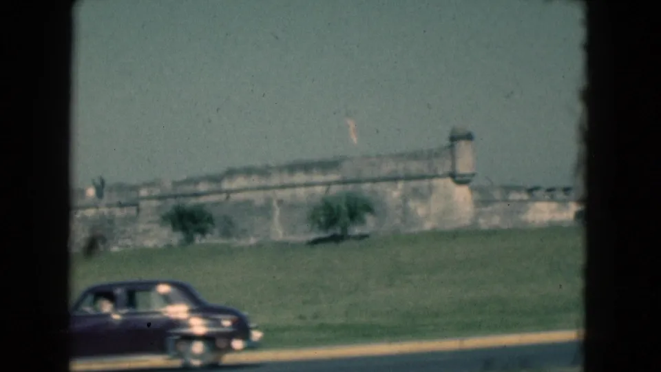 1975 Florida home-movie frame with a dark sedan crossing the foreground below the fort wall, flag, lawn, and trees.